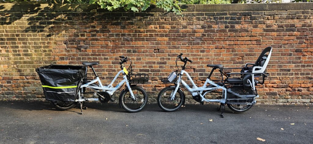 Two white electric longtail cargo bikes, one with a cargo box and one with a child seat, against a sunlit London brick wall